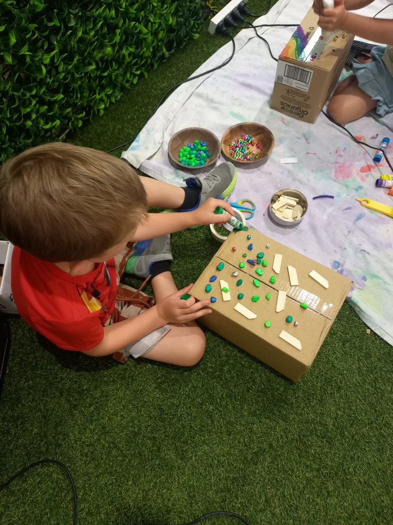 Boy using a green paint stick on a cardboard box he had decorated with green and blue stones and pieces of wood before.